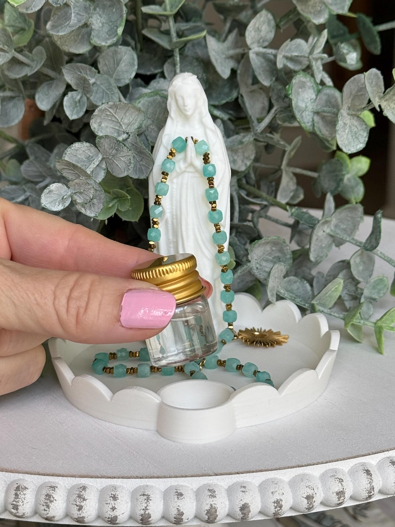 Peut inclure: Un petit flacon en verre transparent avec un bouchon &agrave; vis dor&eacute;, un collier de perles bleu clair &agrave; facettes avec des entretoises dor&eacute;es, et une figurine blanche repr&eacute;sentant une personne en pri&egrave;re, sur un plateau blanc festonn&eacute;.