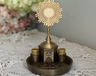 May include: A gold-colored monstrance featuring a central circular sunburst design topped with a small cross. The base is a dark bronze-toned circular tray with a central architectural structure and two small cylindrical containers with gold lids.