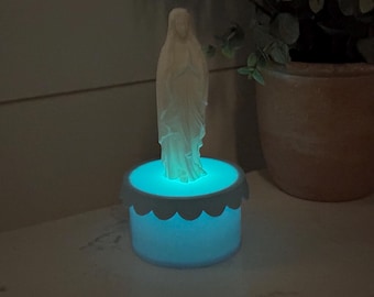 May include: A white statue of a religious figure stands atop a glowing blue base with a scalloped edge. The statue is detailed with flowing robes and a serene expression. The base emits a soft, ambient light.