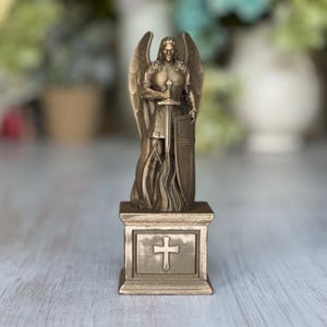 May include: A gold-colored metal statue of an angel warrior holding a sword and shield, standing on a square base with a cross design.