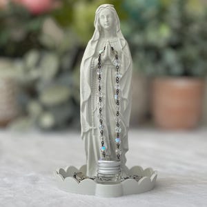 Peut inclure: Une statue blanche mate de la Vierge Marie debout sur une base circulaire festonn&eacute;e. La figure a les mains jointes en pri&egrave;re, v&ecirc;tue d'une longue robe. Un chapelet aux perles transparentes et iris&eacute;es et aux maillons sombres est suspendu.