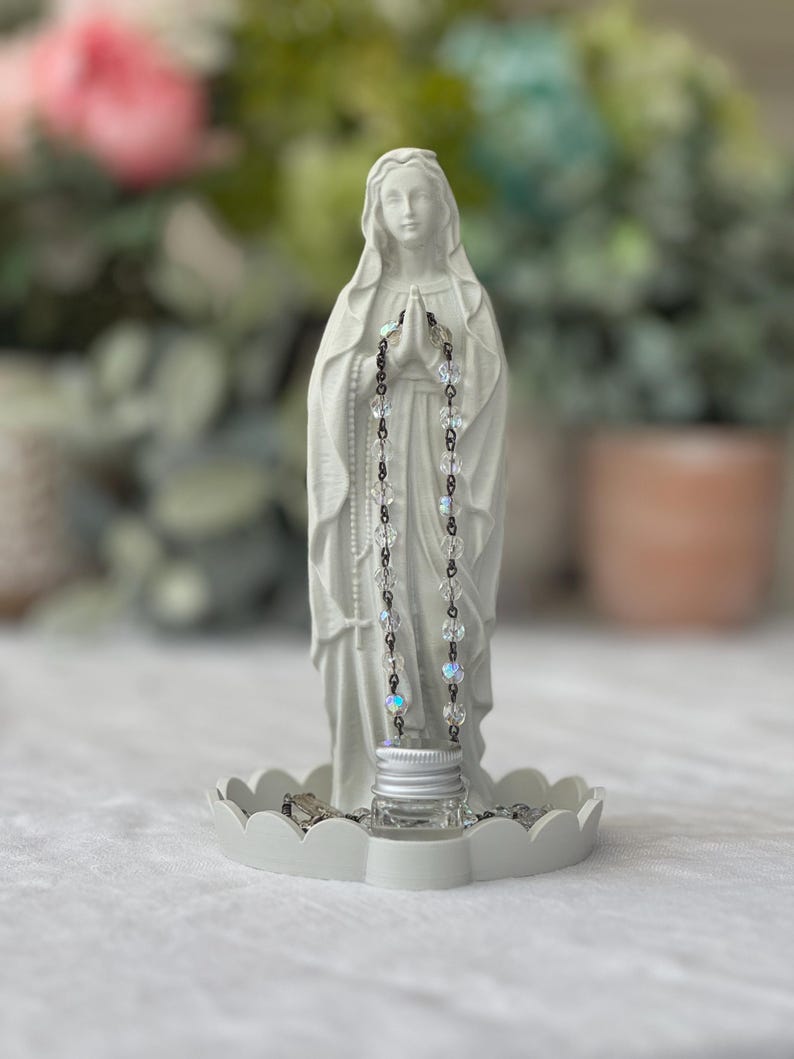 Peut inclure: Une statue blanche mate de la Vierge Marie debout sur une base circulaire festonn&eacute;e. La figure a les mains jointes en pri&egrave;re, v&ecirc;tue d'une longue robe. Un chapelet aux perles transparentes et iris&eacute;es et aux maillons sombres est suspendu.