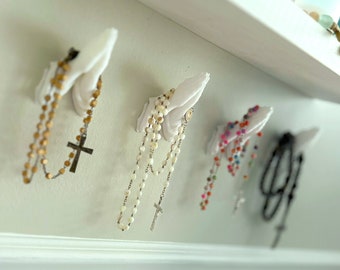 May include: Four white wall-mounted hooks shaped like praying hands, each holding a different rosary. The rosaries feature beads in various colors including brown wood, white pearl, multi-colored glass, and black, each with a metal crucifix.
