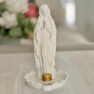 Peut inclure: Une figurine blanche mate repr&eacute;sentant une silhouette aux mains jointes en pri&egrave;re, pos&eacute;e sur une base circulaire festonn&eacute;e. Un petit flacon en verre transparent avec un bouchon &agrave; vis dor&eacute; m&eacute;tallique est plac&eacute; devant la figurine sur la base.
