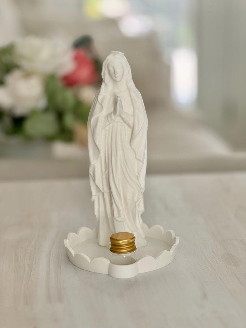 Peut inclure: Une figurine blanche mate repr&eacute;sentant une silhouette aux mains jointes en pri&egrave;re, pos&eacute;e sur une base circulaire festonn&eacute;e. Un petit flacon en verre transparent avec un bouchon &agrave; vis dor&eacute; m&eacute;tallique est plac&eacute; devant la figurine sur la base.