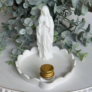 Peut inclure: Une figurine blanche repr&eacute;sentant une personne les mains jointes en pri&egrave;re, plac&eacute;e sur un plateau circulaire blanc &agrave; bord festonn&eacute;. Un petit flacon en verre transparent avec un bouchon &agrave; vis dor&eacute; m&eacute;tallique repose sur le plateau devant la figurine.
