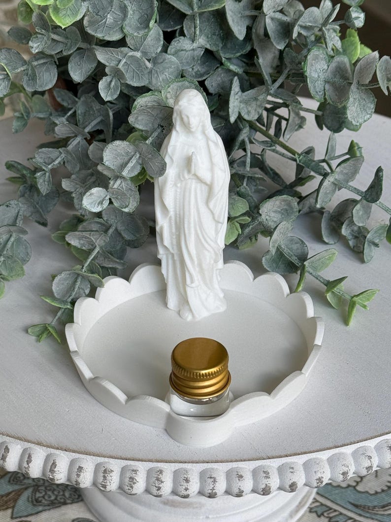 Peut inclure: Une figurine blanche repr&eacute;sentant une personne les mains jointes en pri&egrave;re, plac&eacute;e sur un plateau circulaire blanc &agrave; bord festonn&eacute;. Un petit flacon en verre transparent avec un bouchon &agrave; vis dor&eacute; m&eacute;tallique repose sur le plateau devant la figurine.