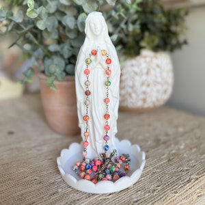 Peut inclure: Une figurine blanche de la Vierge Marie debout sur une base circulaire festonn&eacute;e, avec un chapelet aux perles multicolores marbr&eacute;es drap&eacute; sur les mains et accumul&eacute; &agrave; la base. Le chapelet comprend un crucifix de couleur argent&eacute;e en bas.