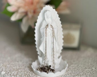 May include: White ceramic figurine of the Virgin Mary with a beaded rosary draped around her neck. The figurine is standing on a white base with a scalloped edge.