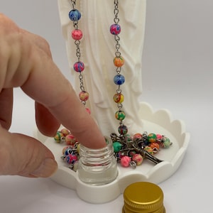 Peut inclure: Une statue blanche mate repr&eacute;sentant une silhouette drap&eacute;e, accompagn&eacute;e d'un chapelet aux perles multicolores marbr&eacute;es et d'un crucifix en m&eacute;tal. Un petit flacon en verre transparent avec un bouchon dor&eacute; est pos&eacute; devant sur une base festonn&eacute;e.