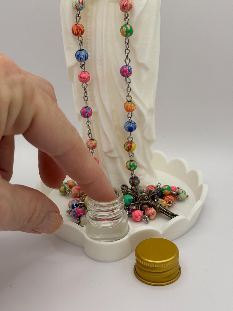 Peut inclure: Une statue blanche mate repr&eacute;sentant une silhouette drap&eacute;e, accompagn&eacute;e d'un chapelet aux perles multicolores marbr&eacute;es et d'un crucifix en m&eacute;tal. Un petit flacon en verre transparent avec un bouchon dor&eacute; est pos&eacute; devant sur une base festonn&eacute;e.