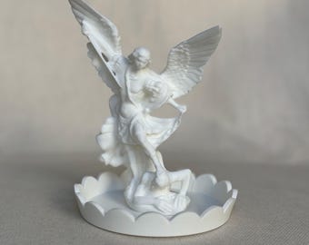 May include: White ceramic sculpture of an angel defeating a demon. The angel has wings and is standing on the demon, who is lying on the ground. The sculpture is on a white base with a scalloped edge.