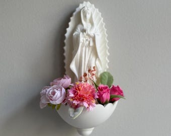 May include: White ceramic wall-mounted holy water font with a sculpted image of the Virgin Mary and a small bouquet of pink and white flowers.