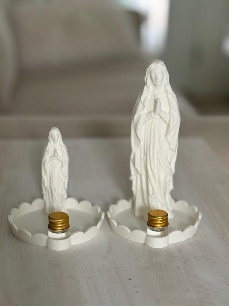 Peut inclure: Deux statues blanches de la Vierge Marie en position de pri&egrave;re, chacune mont&eacute;e sur un plateau circulaire festonn&eacute;. Chaque plateau comporte un petit flacon en verre transparent avec un bouchon &agrave; vis dor&eacute; m&eacute;tallique devant la figurine.