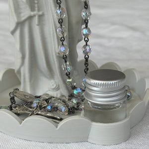 Peut inclure: Un plateau circulaire blanc &agrave; bords festonn&eacute;s supportant une figurine blanche repr&eacute;sentant une personne en robe, un chapelet aux perles transparentes iris&eacute;es avec un crucifix argent&eacute;, et un petit flacon en verre avec un bouchon argent&eacute;.