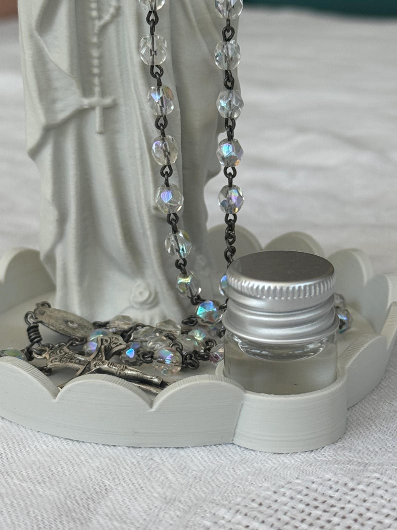 Peut inclure: Un plateau circulaire blanc &agrave; bords festonn&eacute;s supportant une figurine blanche repr&eacute;sentant une personne en robe, un chapelet aux perles transparentes iris&eacute;es avec un crucifix argent&eacute;, et un petit flacon en verre avec un bouchon argent&eacute;.