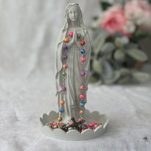 Peut inclure: Une figurine blanc mat repr&eacute;sentant une silhouette aux mains jointes en pri&egrave;re, pos&eacute;e sur une base circulaire festonn&eacute;e. Un chapelet aux perles marbr&eacute;es multicolores et un crucifix en m&eacute;tal sombre sont drap&eacute;s sur les mains et pos&eacute;s sur la base.