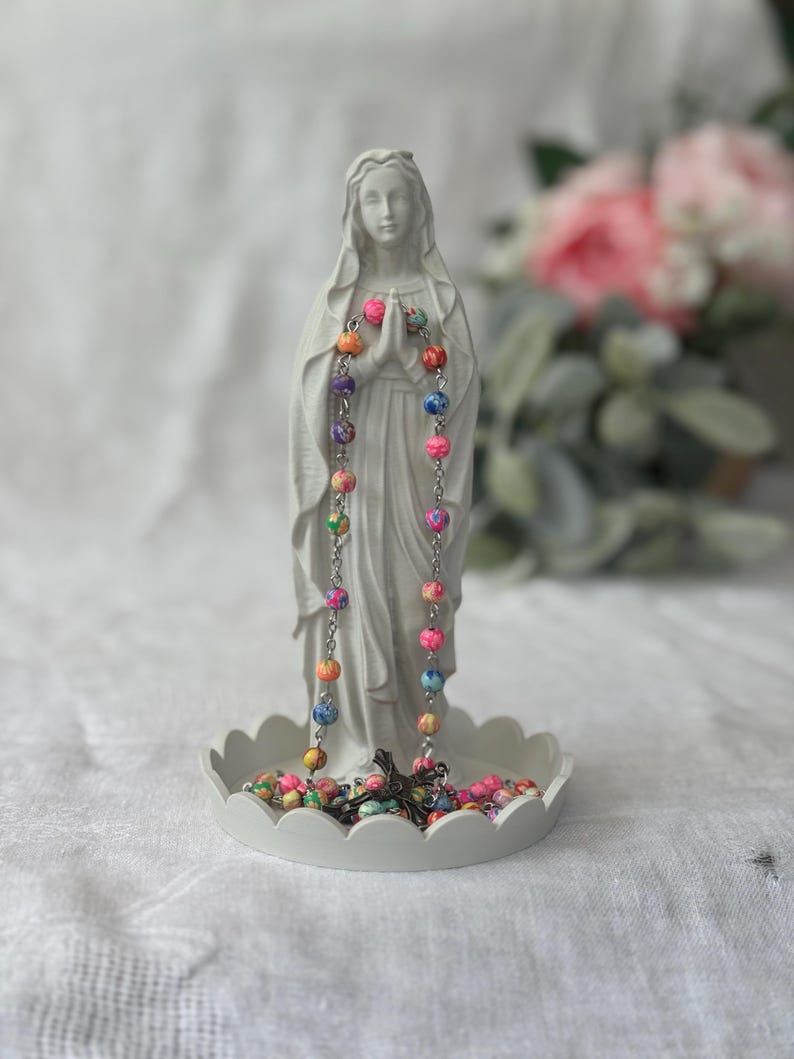 Peut inclure: Une figurine blanc mat repr&eacute;sentant une silhouette aux mains jointes en pri&egrave;re, pos&eacute;e sur une base circulaire festonn&eacute;e. Un chapelet aux perles marbr&eacute;es multicolores et un crucifix en m&eacute;tal sombre sont drap&eacute;s sur les mains et pos&eacute;s sur la base.