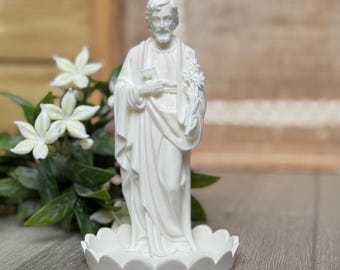 May include: A white statue of a man in flowing robes, holding a key and flowers, stands on a scalloped base. The statue is set against a blurred background with white flowers and greenery.