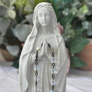 Peut inclure: Une figurine blanc mat de la Vierge Marie avec un voile drap&eacute; sur la t&ecirc;te et les mains jointes en pri&egrave;re. Un chapelet compos&eacute; de perles de verre transparentes, facett&eacute;es et iris&eacute;es, reli&eacute;es par des maillons en m&eacute;tal sombre, est pos&eacute; sur ses mains.