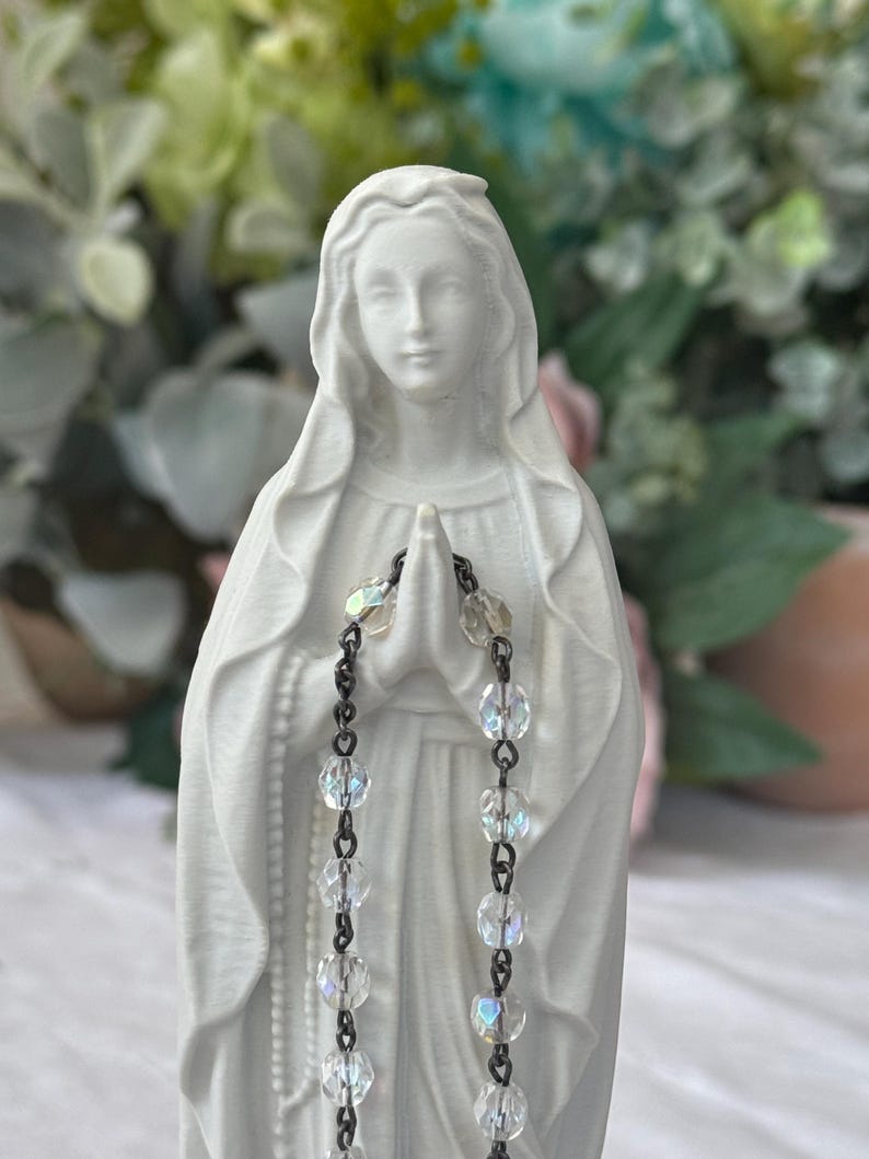 Peut inclure: Une figurine blanc mat de la Vierge Marie avec un voile drap&eacute; sur la t&ecirc;te et les mains jointes en pri&egrave;re. Un chapelet compos&eacute; de perles de verre transparentes, facett&eacute;es et iris&eacute;es, reli&eacute;es par des maillons en m&eacute;tal sombre, est pos&eacute; sur ses mains.