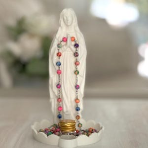 Peut inclure: Une figurine blanche mate repr&eacute;sentant une silhouette debout, les mains jointes en pri&egrave;re, v&ecirc;tue d'une longue robe. Un chapelet de perles multicolores &agrave; motifs floraux est suspendu autour du cou, reposant sur un plateau blanc festonn&eacute;.