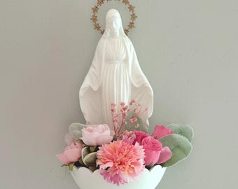 May include: A white wall-mounted basin containing an arrangement of pink and coral artificial flowers and green leaves. A white figurine of a figure in a long robe with a gold-colored star halo stands upright within the floral display.