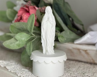 May include: White ceramic figurine of the Virgin Mary standing on a white scalloped base. The figurine is wearing a long white robe and has her hands clasped in prayer.