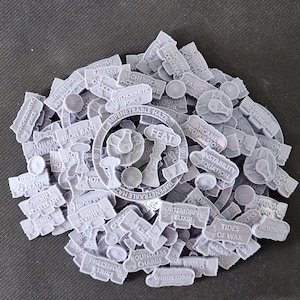 May include: A pile of gray, 3D-printed game tokens with various text inscriptions. The tokens include words like "Feat", "Windstorm", and "Precision Strike". The tokens are scattered on a dark surface.