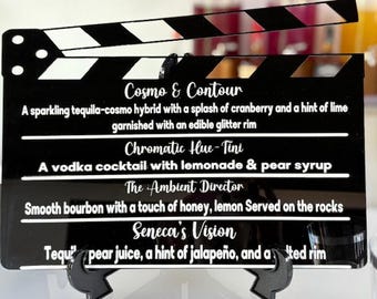 May include: Black acrylic clapperboard with white text listing cocktail names and descriptions. The clapperboard has a black and white striped top section and is displayed on a black stand.