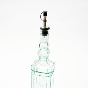 Könnte beinhalten: Eine klare Glasflasche mit strukturierter Oberfläche und einem silbernen Ausgießer. Die Flasche hat eine grüne Tönung und ist für Öl oder Essig gedacht.