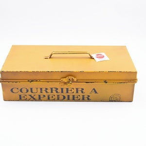 Könnte beinhalten: Senfgelbe Metallwerkzeugkiste mit Griff und Verschluss. Auf der Box steht in schwarzer Schrift "COURRIER A EXPEDIER". Ein kleines Etikett ist oben angebracht.