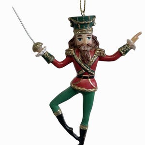 Könnte beinhalten: Dekorative Weihnachtsbaum-Ornament eines Nussknacker-Soldaten in einer Ballettpose. Das Ornament hat eine rote Jacke mit goldenen Verzierungen, grüne Hosen und schwarze Stiefel. Der Nussknacker hält ein Schwert und trägt einen grünen Hut.