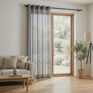 Può includere: Una tenda grigia semi-trasparente a tutta altezza pende da un bastone in metallo scuro sopra una porta a vetri. La tenda presenta occhielli metallici distanziati che creano pieghe verticali, permettendo alla luce di filtrare attraverso.