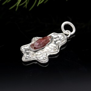 May include: A silver pendant featuring a raw, reddish-brown gemstone set in a hammered, star-shaped frame. The pendant has a textured surface and a small loop for a chain. The background is black.