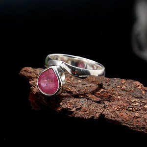 Puede incluir: Un anillo de plata con una gema rosa en forma de pera. La banda del anillo tiene un dise&ntilde;o curvo &uacute;nico. La gema est&aacute; engastada en un bisel y el anillo se muestra sobre madera texturizada sobre un fondo negro.