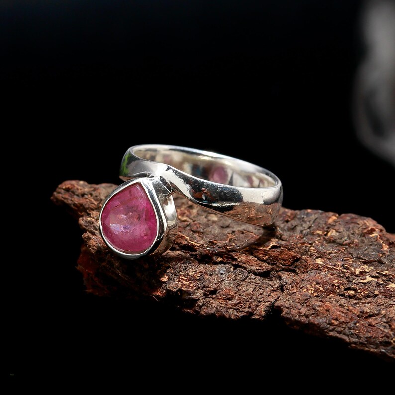 Puede incluir: Un anillo de plata con una gema rosa en forma de pera. La banda del anillo tiene un dise&ntilde;o curvo &uacute;nico. La gema est&aacute; engastada en un bisel y el anillo se muestra sobre madera texturizada sobre un fondo negro.
