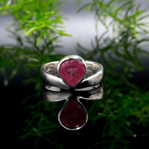 Puede incluir: Un anillo de plata con una gema rosa en forma de pera. El anillo est&aacute; sobre una superficie negra reflectante, con follaje verde en el fondo. La gema es de color rosa vibrante.