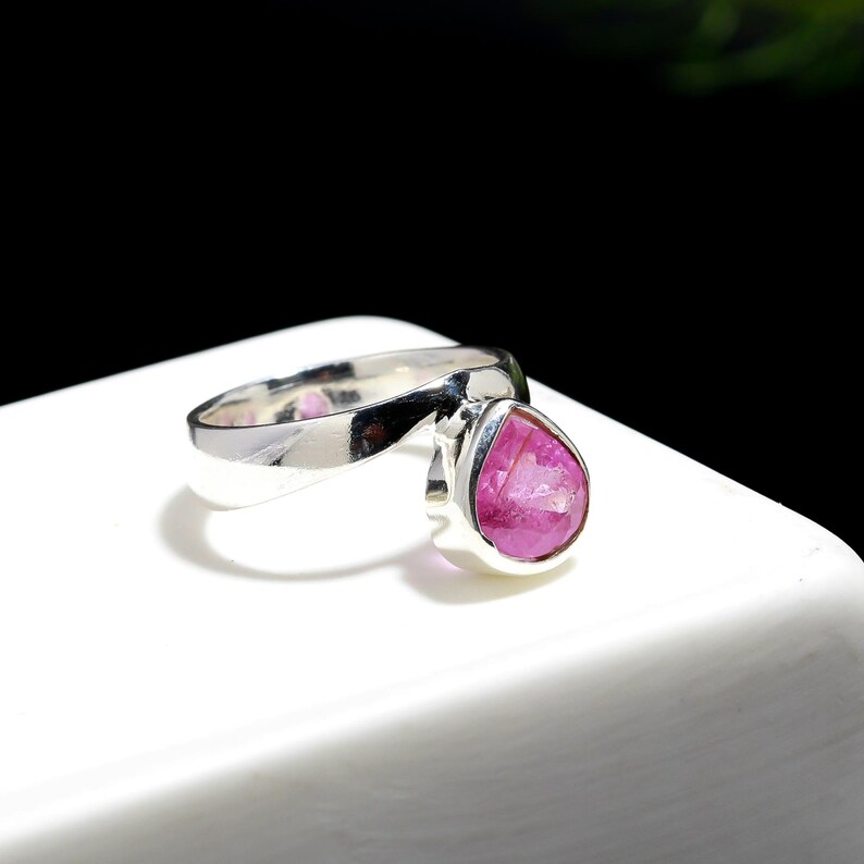 Puede incluir: Un anillo de plata con una piedra preciosa rosa en forma de pera. La banda del anillo se curva elegantemente, conduciendo al engaste que sostiene la vibrante piedra. El color de la piedra es un rosa rico y saturado.