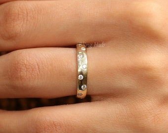 May include: A gold band ring featuring a series of small, round, clear stones set flush into the metal surface. The ring has a smooth, polished finish and is worn on a finger, displaying the circular arrangement of the embedded stones clearly.