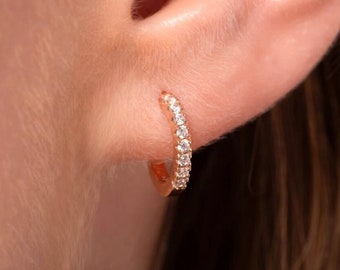 May include: A small rose gold hoop earring featuring a row of clear, sparkling stones embedded along the front curve. The circular metal band has a polished finish and is secured through the earlobe, showcasing a delicate and refined design.