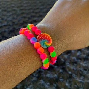 May include: Two colourful beaded bracelets with a rainbow swirl charm. The bracelets are made of pink, orange, yellow, green, and blue beads.