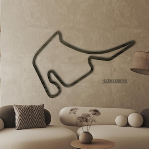 Puede incluir: Un contorno oscuro de un circuito de carreras, montado en una pared sobre un sofá color crema. La palabra "Hockenheimring" está debajo. Una pantalla de lámpara tejida cuelga de una lámpara curva. La habitación tiene una paleta de colores neutros.