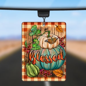 May include: A car air freshener with a checkered background and a blue and white pumpkin with the word "Blessed" on it. There is a butterfly on the top pumpkin and a small green pumpkin on the bottom.