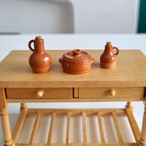May include: Miniature terracotta kitchen set on a wooden table. The set includes a small pot with a lid, and two jugs with cork stoppers. The table has two drawers and a lower shelf. The set is likely for a dollhouse or decorative display.