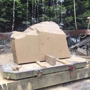 May include: A large, beige, rough-hewn stone block sits on a metal industrial cutting platform. Water streams down from above, cooling the surface of the stone as it is being processed by a heavy-duty saw blade, creating a wet, textured appearance.