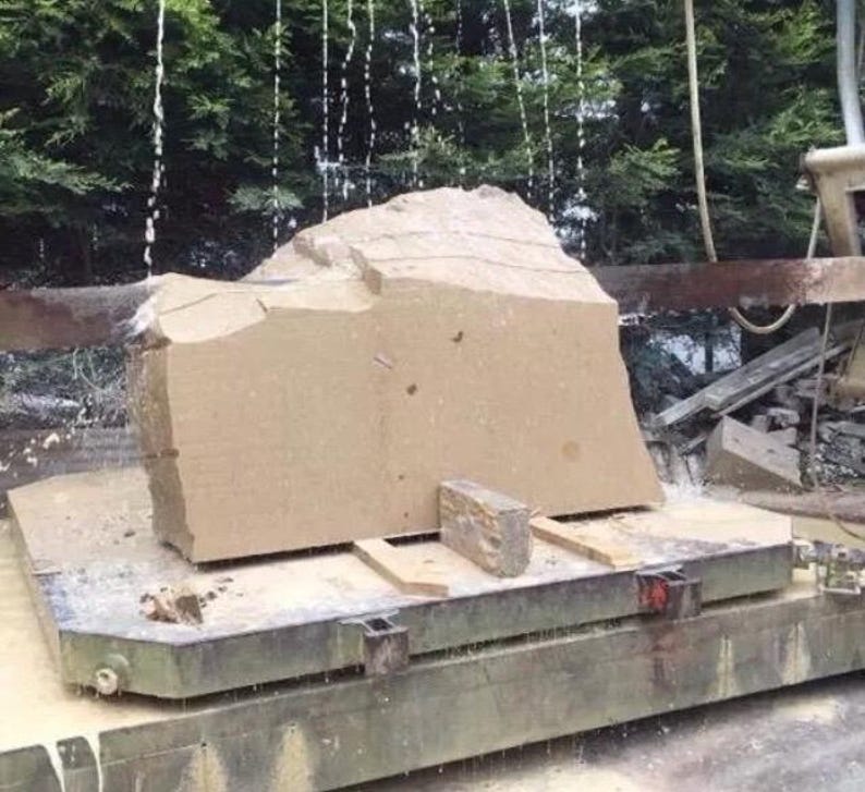 May include: A large, beige, rough-hewn stone block sits on a metal industrial cutting platform. Water streams down from above, cooling the surface of the stone as it is being processed by a heavy-duty saw blade, creating a wet, textured appearance.
