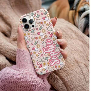 May include: A pink phone case featuring a pattern of medical symbols, including stethoscopes, bandages, pills, and syringes. The name "Sophie" is written in cursive script. The case is designed for a smartphone.