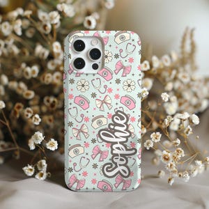 May include: A light green phone case with a pattern of pink bows, nurse hats, stethoscopes, and flowers. The name "Sophie" is written in cursive. The case has a camera cutout and is surrounded by white flowers.