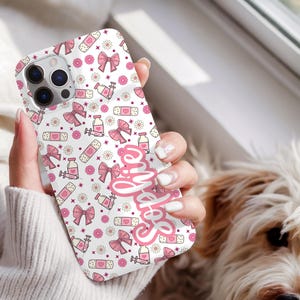 May include: A white phone case with a pink pattern of bows, bandages, flowers, and syringes. The name "Sophie" is written in pink script on the case. The case is held in a hand.